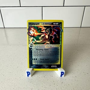 Peut inclure: Une carte à collectionner Pokémon Charizard, présentée dans un support blanc avec la lettre bleue "P" de chaque côté. La carte présente une illustration de dragon de feu avec le texte "Charizard" et "90 HP".