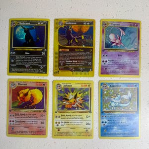 May include: A collection of six rectangular trading cards featuring illustrations of Umbreon, Espeon, Flareon, Jolteon, and Vaporeon. Each card displays character names, HP values, attack descriptions, and game statistics printed in black text on colored backgrounds.