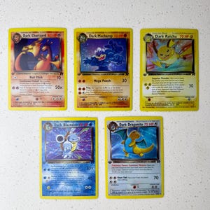 Puede incluir: Cinco cartas coleccionables de Pokémon con Dark Charizard, Dark Machamp, Dark Raichu, Dark Blastoise y Dark Dragonite. Las cartas tienen bordes amarillos e ilustraciones coloridas de los Pokémon. Cada carta muestra el nombre, los HP y los detalles de ataque del Pokémon.