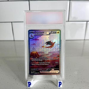 May include: A Charizard EX Pokémon trading card, graded and encased in a clear plastic holder. The card features a holographic image of a fiery dragon over a volcanic landscape. The card is displayed on a white stand.