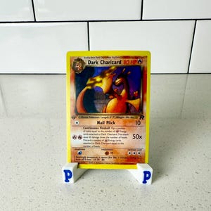 Könnte beinhalten: Eine gelbe Dark Charizard Pokémon-Sammelkarte mit einer Illustration eines orangefarbenen und schwarzen Drachen. Die Karte wird auf einem weißen Ständer mit dem Buchstaben "P" auf jeder Seite angezeigt. Der Kartentext enthält "Nail Flick" und "Continuous Fireball".