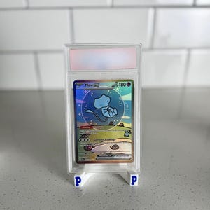 May include: A graded Mew ex Pokémon trading card in a clear protective case. The card features a holographic design with a blue Mew character. The card is displayed on a white stand with blue "P" logos.