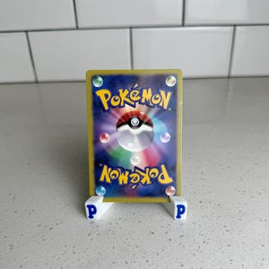 Pode incluir: Um cart&atilde;o colecion&aacute;vel Pok&eacute;mon exibido em um suporte branco com letras "P" azuis. A parte de tr&aacute;s do cart&atilde;o apresenta o logotipo Pok&eacute;mon, uma Pok&eacute; Bola e orbes coloridos em um fundo azul escuro. O cart&atilde;o tem uma borda dourada.