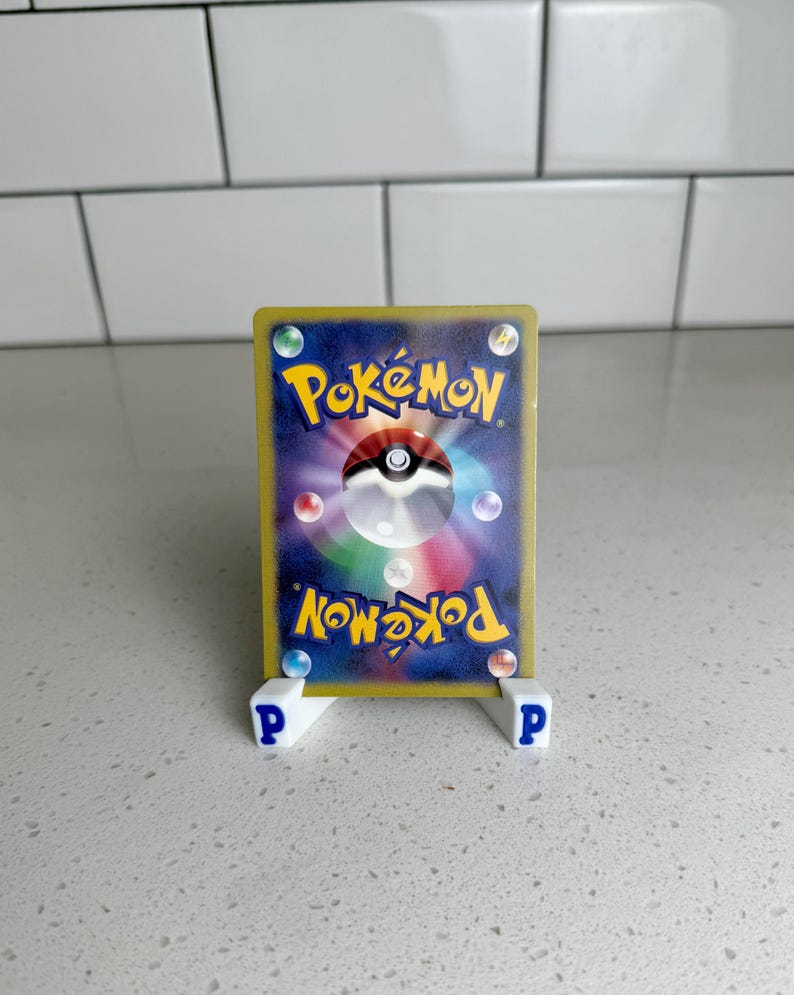 Pode incluir: Um cart&atilde;o colecion&aacute;vel Pok&eacute;mon exibido em um suporte branco com letras "P" azuis. A parte de tr&aacute;s do cart&atilde;o apresenta o logotipo Pok&eacute;mon, uma Pok&eacute; Bola e orbes coloridos em um fundo azul escuro. O cart&atilde;o tem uma borda dourada.