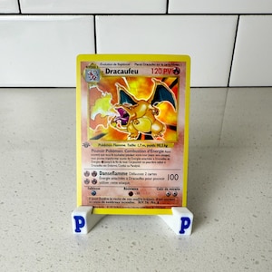 Könnte beinhalten: Gelbe Dracaufeu Pokémon-Sammelkarte mit einer Illustration von Charizard mit orangefarbenen und schwarzen Flügeln. Die Karte wird auf einem weiß-blauen Ständer präsentiert. Text enthält "Dracaufeu" und "120 PV".