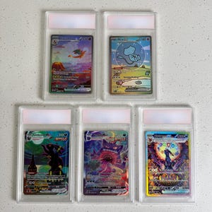 May include: Five Pokémon trading cards in protective cases. The cards feature various characters and colourful designs. The cards are in clear plastic holders with white tops, arranged on a white surface.