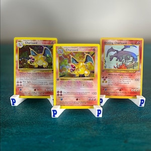 May include: Three collectible trading cards featuring Charizard and Shining Charizard characters, each displayed on a small white plastic stand with a blue letter P printed on the front. The cards display detailed illustrations, text, and game statistics.