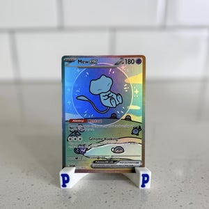 May include: A holographic Mew EX Pokémon trading card displayed in a white stand. The card features a blue Mew character with the text "Mew EX" and "180". The card is set against a bright background.