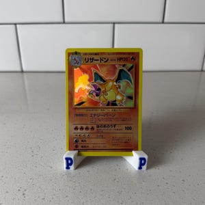 Puede incluir: Una tarjeta coleccionable Pokémon Charizard amarilla, exhibida en un soporte blanco con logotipos "P" azules. La tarjeta presenta una ilustración ardiente de Charizard y texto japonés.