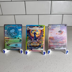 May include: Three Pokémon trading cards on white stands with the letter "P". The cards show colourful illustrations of Pokémon characters. The cards are arranged in a row against a white tiled background.