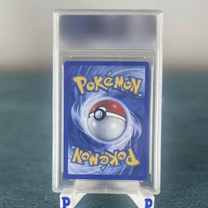 May include: A graded Pok&eacute;mon trading card, encased in a clear plastic holder. The card features a blue background with the Pok&eacute;mon logo and a Pok&eacute; Ball design. The holder is supported by a white stand with the letter "P" on each side.