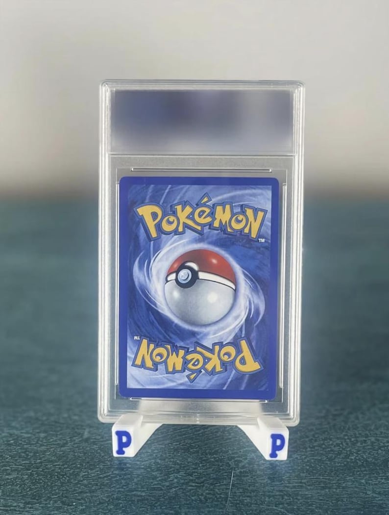 May include: A graded Pok&eacute;mon trading card, encased in a clear plastic holder. The card features a blue background with the Pok&eacute;mon logo and a Pok&eacute; Ball design. The holder is supported by a white stand with the letter "P" on each side.