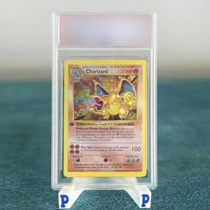 以下が含まれることがあります： 透明な保護ケースに入った、評価済みのリザードンポケモンカード。カードには、「120 HP」や「Fire Spin」などのテキストとともに、黄色とオレンジ色で描かれたリザードンのイラストが描かれています。カードは、青色の「P」ロゴが付いた白いスタンドに表示されています。