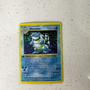 May include: A holographic Blastoise trading card with a yellow border, featuring the character in the center, 100 HP, the Rain Dance Pokémon Power, and the Hydro Pump attack. The card text includes details like Shellfish Pokémon, 5' 3" length, and 189 lbs weight.