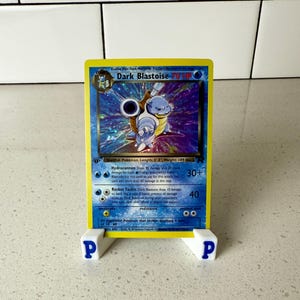 Puede incluir: Una carta coleccionable de Pokémon Dark Blastoise, con una criatura azul con forma de tortuga y un borde amarillo. La carta se muestra en un soporte blanco con una "P" azul a cada lado. El texto de la carta incluye "Hydrocannon" y "Rocket Tackle".