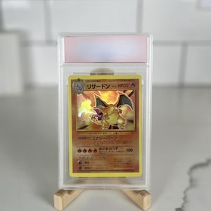 Puede incluir: Una carta Pokémon Charizard graduada en una funda protectora transparente. La carta presenta una ilustración dorada y naranja de Charizard con texto japonés. La carta se muestra sobre un soporte de madera sobre un fondo blanco.