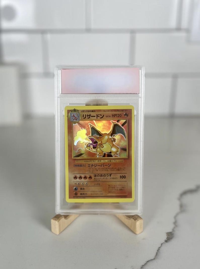 Op de afbeelding: Een beoordeelde Charizard Pok&eacute;mon-kaart in een doorzichtige beschermhoes. De kaart toont een goud-oranje illustratie van Charizard met Japanse tekst. De kaart wordt gepresenteerd op een houten standaard tegen een witte achtergrond.