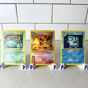 May include: Three vintage Pokémon trading cards: Venusaur, Charizard, and Blastoise. Each card is displayed on a white stand with a blue "P" on the front. The cards have yellow borders and feature colorful illustrations of the Pokémon.