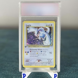 Puede incluir: Una carta coleccionable de Pokémon calificada con Lugia, un Pokémon Buceo. La carta está encerrada en un soporte de plástico transparente y se muestra en un soporte blanco con la letra "P" a cada lado. La tarjeta tiene una ilustración azul y blanca.