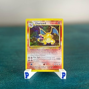 May include: A Charizard trading card featuring a yellow border, a fire-type illustration, and 120 HP, displayed on a white plastic stand with a blue letter P on each side. The card text includes details like Energy Burn, Fire Spin, and 100 damage.