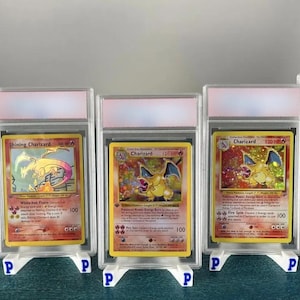May include: Three graded Pok&eacute;mon trading cards featuring Charizard. The cards are encased in clear plastic holders and displayed on white stands. The cards have colorful illustrations and text, including "Shining Charizard" and "Charizard".