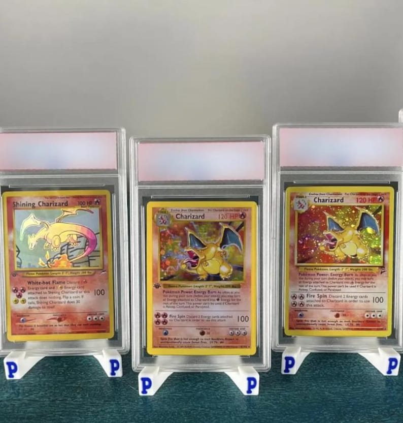 May include: Three graded Pok&eacute;mon trading cards featuring Charizard. The cards are encased in clear plastic holders and displayed on white stands. The cards have colorful illustrations and text, including "Shining Charizard" and "Charizard".