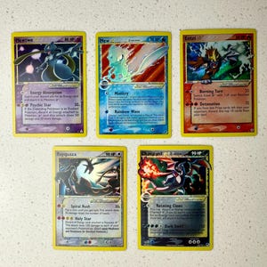 May include: Five Pokémon trading cards are displayed, featuring characters like Mewtwo, Mew, Entei, Rayquaza, and Charizard. Each card has a unique design with character art, HP, and attack descriptions. The cards are on a light-colored surface.