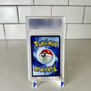 Op de afbeelding: Een beoordeelde Pok&eacute;mon-verzamelkaart in een doorzichtige plastic hoes. De kaart toont het Pok&eacute;mon-logo en een Pok&eacute; Ball-ontwerp op een blauwe achtergrond. De hoes wordt ondersteund door twee kleine witte standaards met blauwe "P" letters.