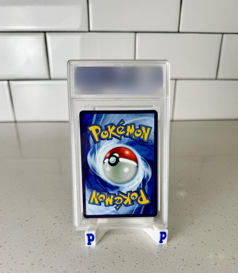 Op de afbeelding: Een beoordeelde Pok&eacute;mon-verzamelkaart in een doorzichtige plastic hoes. De kaart toont het Pok&eacute;mon-logo en een Pok&eacute; Ball-ontwerp op een blauwe achtergrond. De hoes wordt ondersteund door twee kleine witte standaards met blauwe "P" letters.