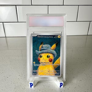 May include: A graded Pokémon trading card featuring Pikachu wearing a blue hat, titled "Pika-Portrait." The card is encased in a clear plastic holder and displayed on a white stand with blue "P" logos.