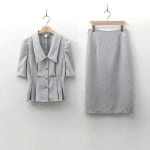 May include: A light gray skirt suit, consisting of a short-sleeved jacket and a knee-length skirt. The jacket has a wide collar, decorative buttons, and a pleated peplum. The skirt is a straight, pencil style. Both pieces are hanging on separate hangers.