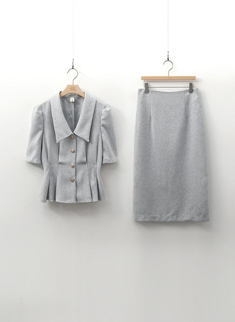 May include: A light gray skirt suit, consisting of a short-sleeved jacket and a knee-length skirt. The jacket has a wide collar, decorative buttons, and a pleated peplum. The skirt is a straight, pencil style. Both pieces are hanging on separate hangers.