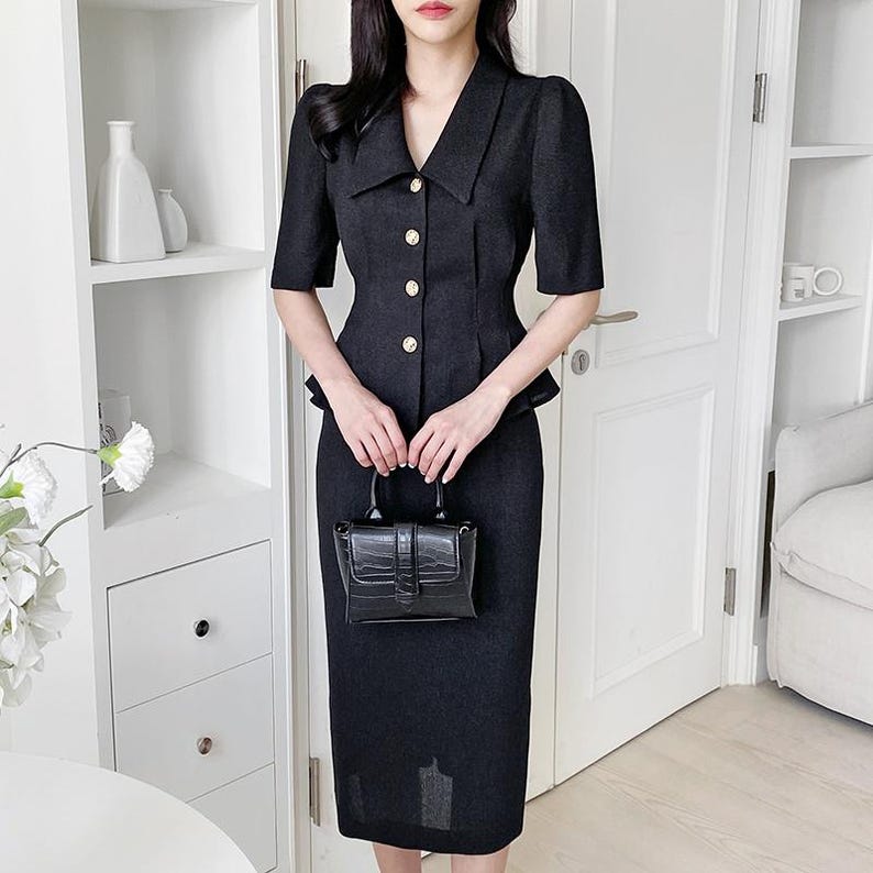 May include: A black two-piece suit featuring a short-sleeved jacket with gold buttons and a matching knee-length pencil skirt. The jacket has a notched collar and a peplum detail. A small black handbag is held in front.