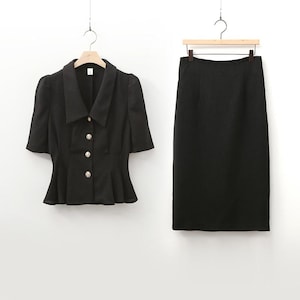 May include: A black two-piece suit, consisting of a short-sleeved jacket with gold buttons and a matching pencil skirt. The jacket has a peplum design and a wide collar. Both pieces are hanging on separate hangers.