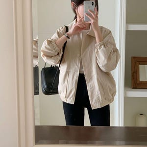 May include: A beige jacket with a button closure and gathered sleeves. The jacket is paired with black pants. A black purse hangs from the shoulder. The image is taken in front of a mirror.