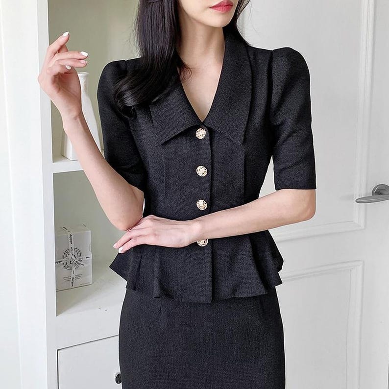 May include: Black two-piece suit featuring a short-sleeved blazer and a matching skirt. The blazer has a V-neck, a peplum waist, and is fastened with three gold-colored buttons. The suit is made of a textured fabric.