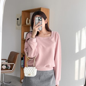 May include: A light pink long-sleeve top with a square neckline and a gray skirt. A small white quilted handbag with a gold chain strap hangs from the shoulder. The outfit is captured in a mirror.