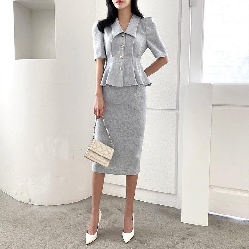 May include: A light gray skirt suit featuring a short-sleeved jacket with gold buttons and a peplum waist, paired with a matching knee-length pencil skirt. The outfit is accessorized with a cream-colored quilted handbag and white pointed-toe heels.