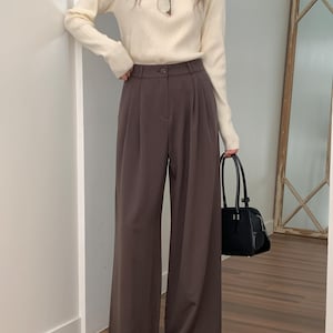 May include: A cream-colored long-sleeve sweater paired with high-waisted, wide-leg brown trousers. A black handbag is held in the hand. The outfit is completed with black shoes and eyeglasses.