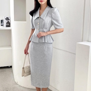 May include: A light gray two-piece suit featuring a short-sleeved jacket with gold buttons and a peplum waist, paired with a matching pencil skirt. The suit is made of a textured fabric and is suitable for business or formal wear. A cream-colored handbag completes the outfit.