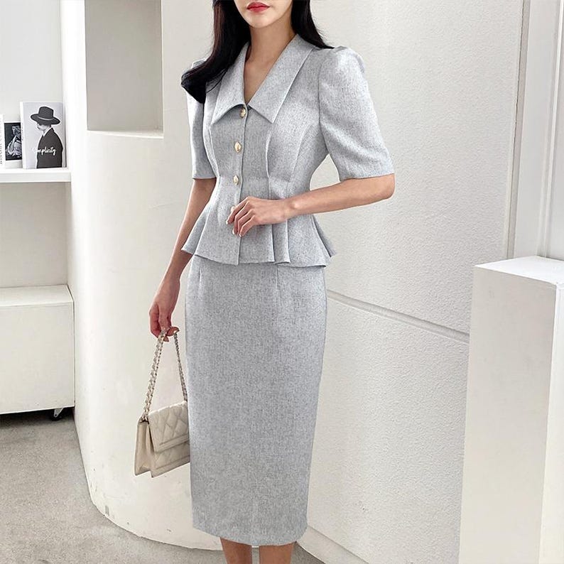 May include: A light gray two-piece suit featuring a short-sleeved jacket with gold buttons and a peplum waist, paired with a matching pencil skirt. The suit is made of a textured fabric and is suitable for business or formal wear. A cream-colored handbag completes the outfit.