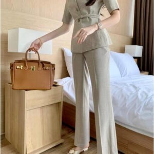 Puede incluir: Un traje de mujer marrón claro compuesto por una blazer de manga corta con cinturón y pantalones de pierna ancha a juego. Un bolso de mano de cuero marrón descansa sobre una mesita de noche de madera. El conjunto se completa con zapatos sin cordones color crema.