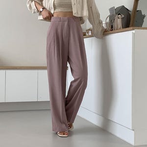 May include: A woman wearing a cream-colored cardigan, a beige crop top, and mauve-colored wide-leg trousers. The trousers have a high waist and pleats. The outfit is paired with white sandals. The background is a minimalist white interior.