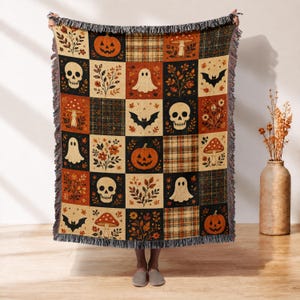 May include: A woven throw blanket featuring a grid pattern of autumn-themed illustrations including orange pumpkins, white skulls, white ghosts, black bats, red mushrooms, and floral foliage in shades of orange, brown, black, and cream with fringe.
