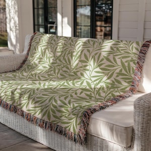 May include: A woven throw blanket with a cream background and a repeating pattern of green leaves and vines. The blanket is draped over a white wicker sofa. The edges are finished with a fringed border in shades of brown, orange, and blue.