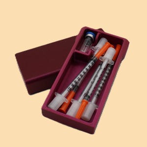 May include: A maroon medical kit containing three syringes with orange caps and clear barrels, a small vial, and a blue-capped bottle. The kit is open, revealing the contents, and sits on a light beige background.