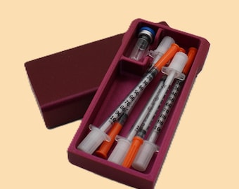 May include: A maroon medical kit containing three syringes with orange caps and clear barrels, a small vial, and a blue-capped bottle. The kit is open, revealing the contents, and sits on a light beige background.