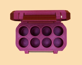 May include: A magenta-colored, open, rectangular case with eight circular compartments. The case has a hinged lid and a silver-colored latch. The case is designed to hold small items, such as makeup or other accessories.