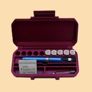 Op de afbeelding: Een pruimkleurige medische reiskoffer, open om een blauwe medische pen, verschillende wit afgedekte flesjes en een alcoholdoekje te onthullen. De koffer heeft een handvat en meerdere compartimenten.