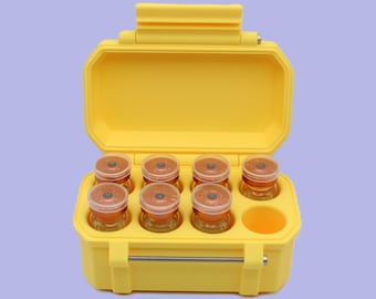 May include: A bright yellow, open, hard-shell case containing eight small glass jars with orange contents and brown lids. The case has a handle and a molded interior to hold the jars securely. The background is a solid light purple.